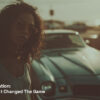 Midjourney Video AI generator A hyper-realistic vintage 1980s muscle car parked at a sunlit beach during golden hour, metallic paint gleaming, soft lens flare, retro surfboards in the back, an 80s-inspired woman with big curly hair, pink sunglasses, denim shorts and a playful smile getting into the car, looking at the camera, cinematic tones, nostalgic color grading, beach vibes